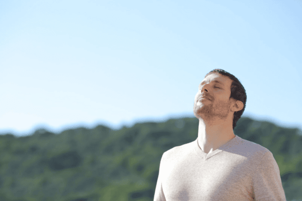 Man standing outdoors with eyes closed, taking a deep breath and enjoying the fresh air under a clear blue sky.